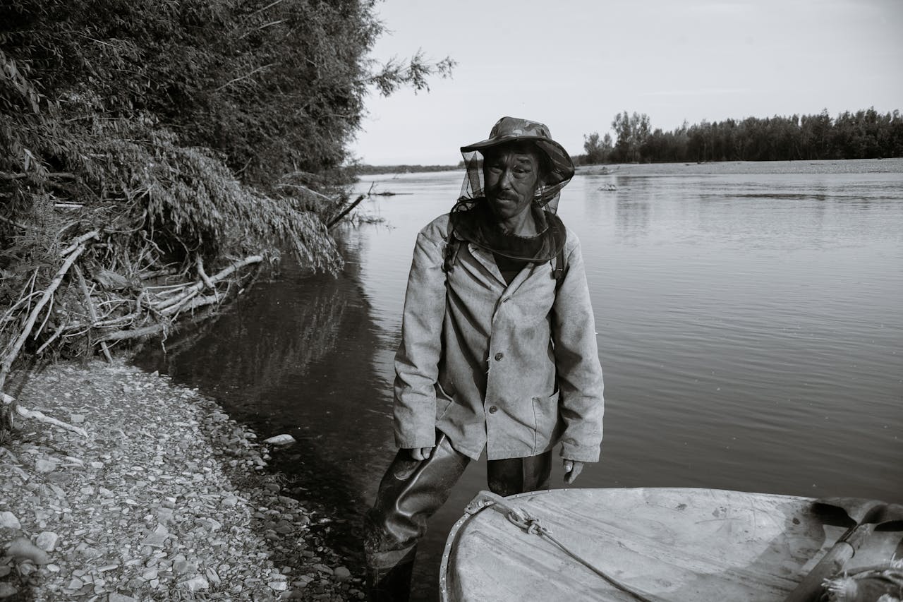 Black and white of calm mature ethnic fisherman in anti mosquito head net hat standing in shallow water of lake near wooden boat and looking at camera